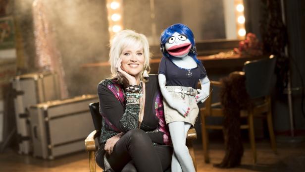 Eine Frau sitzt neben einer Puppe mit blauen Haaren vor einem Schminktisch.