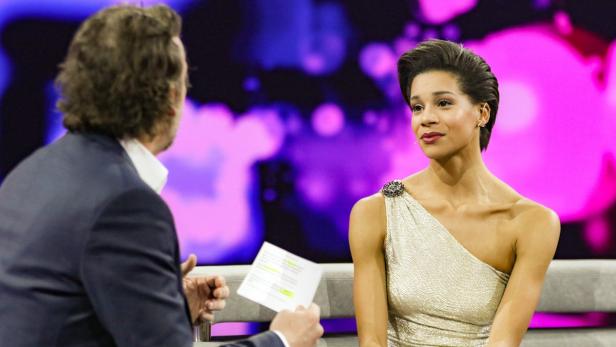 Eine Frau in einem goldenen Kleid sitzt während einer Talkshow auf einem Sofa.