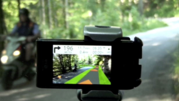 Ein Smartphone zeigt eine Navigations-App, während ein Roller auf einer Straße fährt.
