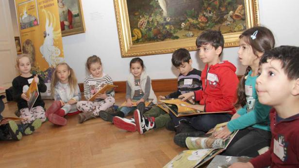 Kinder aus dem Wiener Kindergarten in der Argentinier Straße mit Büchern vor jenem Gemälde von Johann Knapp, in dem sich ein Kakadu befindet.