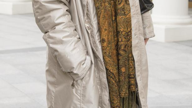 Eine Frau mit braunen Haaren, bekleidet mit einem beigen Mantel und einem gemusterten Schal.