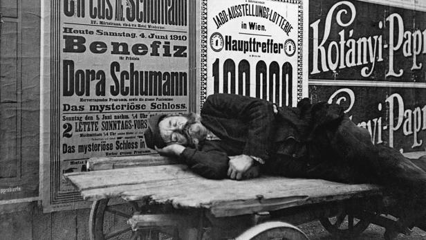 Ein Mann schläft auf einem Karren vor Plakaten in Wien, darunter Werbung für Dora Schumann und eine Jagd-Ausstellungs-Lotterie.