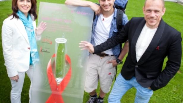 Drei Personen posieren mit einem Transparent, das eine Gurke in einem Kondom zeigt, umgeben von einer roten Schleife.