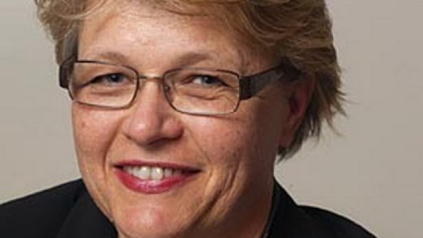 Eine Frau mit blonden Haaren, Brille und dunklem Blazer lächelt in die Kamera.