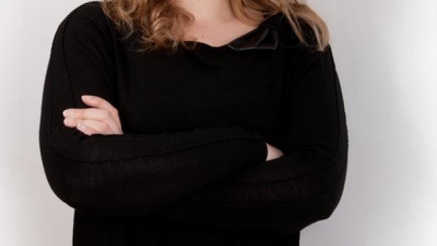 Eine Frau mit blonden Locken lächelt mit verschränkten Armen in die Kamera.
