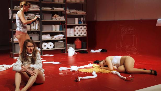 Auf einer roten Bühne liegen und sitzen drei Frauen vor einem Regal und einer Ofenzeichnung.