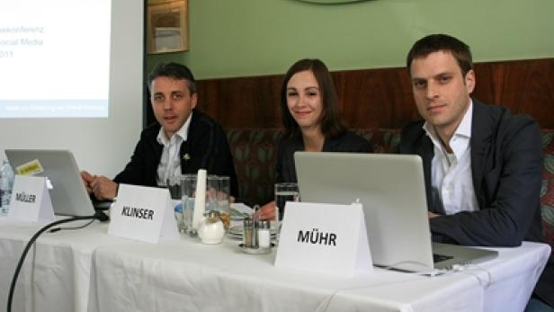 Drei Personen sitzen an einem Tisch mit Laptops und Namensschildern bei einer Pressekonferenz.