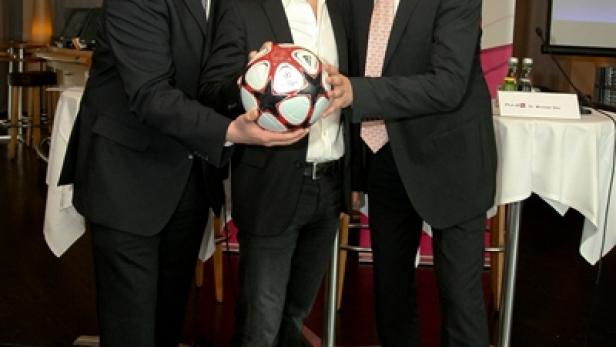 Drei Männer in Anzügen posieren mit einem Fußball in einem Innenraum.