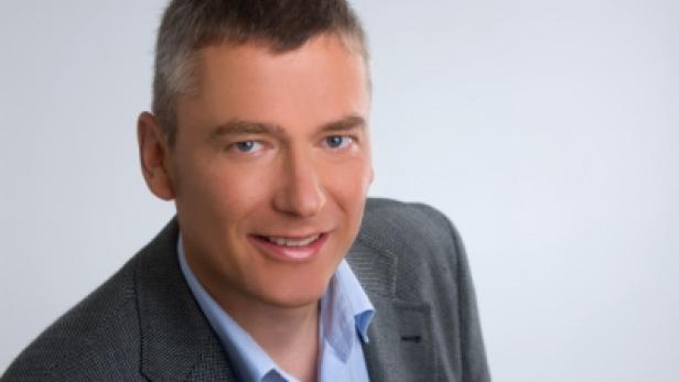 Ein Mann mit grau meliertem Haar lächelt in einem blauen Hemd und einem grauen Sakko.