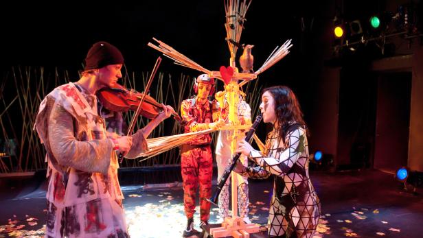 Drei Darsteller musizieren auf einer Bühne mit Geige, Klarinette und einem hölzernen Objekt mit Vogel.