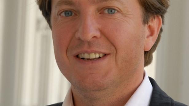 Ein lächelnder Mann mit blonden Haaren und blauem Hemd in einem dunklen Anzug.