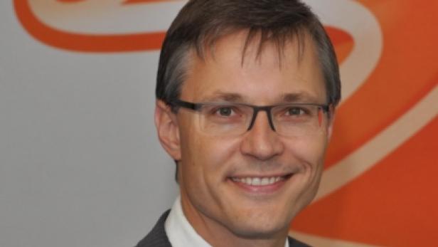 Ein lächelnder Mann mit Brille und Anzug vor einem orangefarbenen Hintergrund.