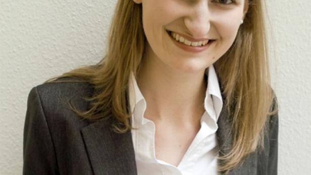 Eine lächelnde Frau in Blazer und weißem Hemd vor einer hellen Wand.
