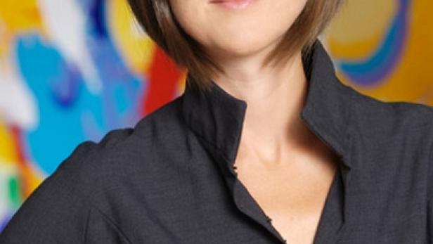 Eine Frau mit braunen Haaren und einem dunklen Blazer vor einem farbenfrohen Hintergrund.