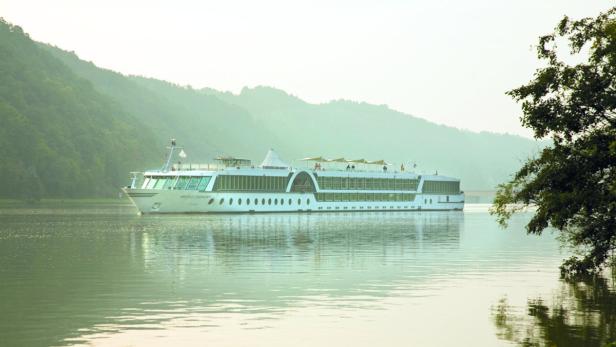 Ein Flusskreuzfahrtschiff fährt auf einem ruhigen Fluss entlang bewaldeter Hügel.