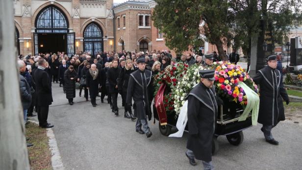 Ein Trauerzug mit einem von Blumen geschmückten Sarg verlässt eine Kirche.