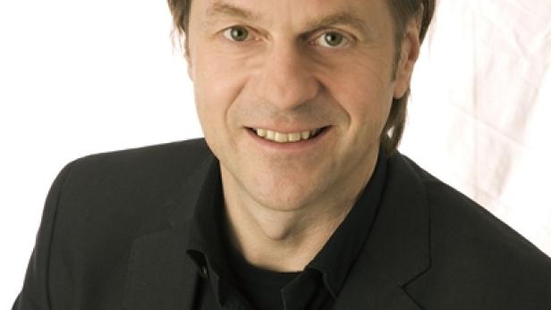 Ein lächelnder Mann in einem schwarzen Anzug vor einem weißen Hintergrund.