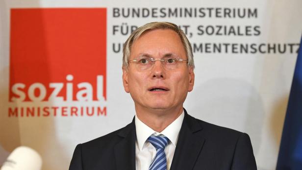 Ein Mann mit Brille und Anzug steht vor dem Logo des Sozialministeriums.