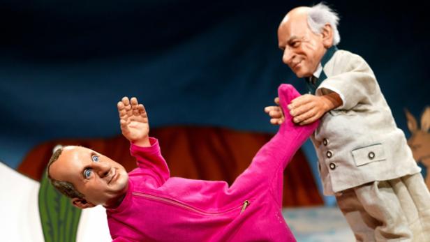 Zwei Marionetten auf einer Bühne, eine in einem pinkfarbenen Overall, die andere in einem Anzug.