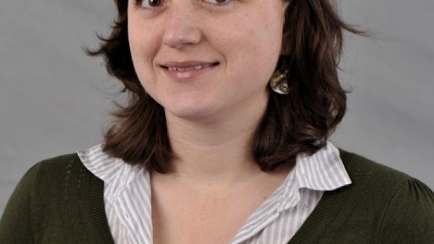 Eine lächelnde Frau mit braunen Haaren und einem grünen Pullover vor einem grauen Hintergrund.