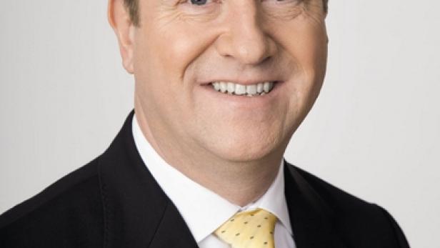 Ein lächelnder Mann im Anzug mit gelber Krawatte vor einem hellen Hintergrund.
