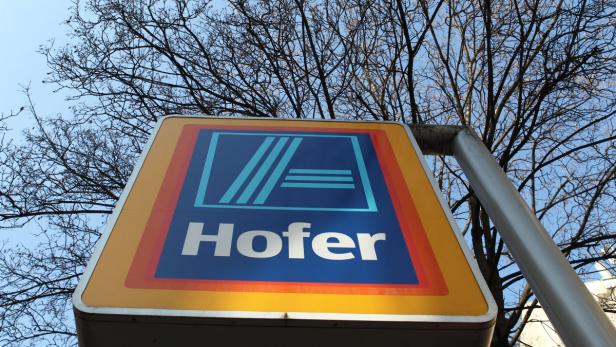 Das Schild einer Hofer-Filiale vor einem Baum und blauem Himmel.