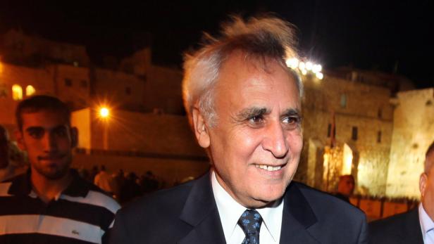 Ein lächelnder Mann im Anzug vor einem beleuchteten Gebäude bei Nacht.