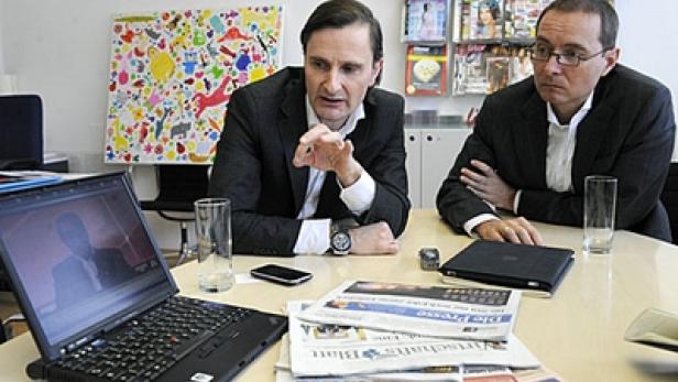 Zwei Männer in Anzügen sitzen an einem Tisch mit Zeitungen und einem Laptop.