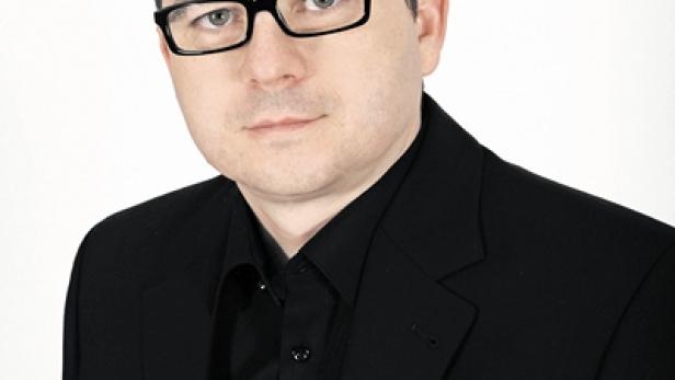 Ein Mann mit Brille und dunklem Anzug vor einem weißen Hintergrund.