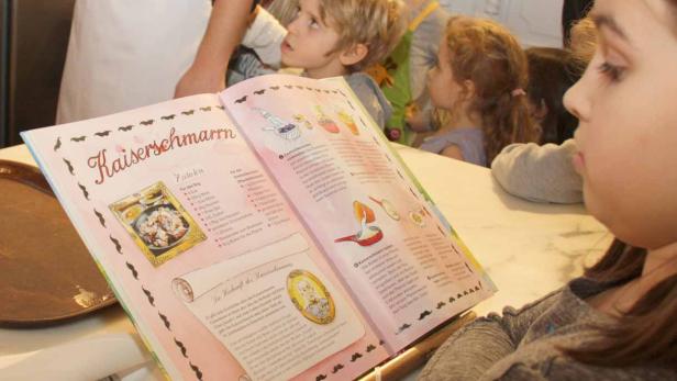 Ein Mädchen liest ein Kochbuch mit dem Rezept für Kaiserschmarrn, während andere Kinder zusehen.