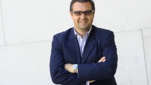 Ein Mann mit Brille und blauem Sakko steht mit verschränkten Armen vor einer Wand.