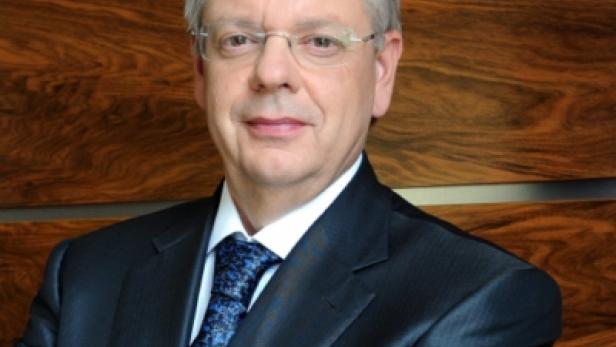 Ein Mann mit Brille und Anzug vor einem Holzhintergrund.