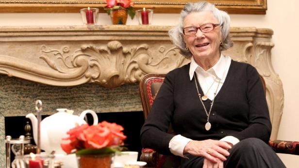 Eine ältere Frau sitzt in einem Sessel vor einem Kamin und einem Teegedeck.