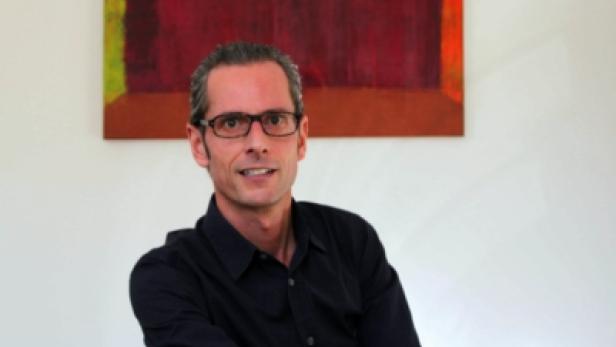 Ein Mann mit Brille und dunklem Hemd vor einem abstrakten Gemälde.