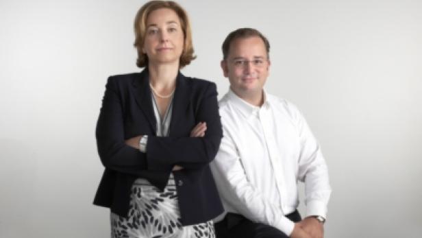 Eine Frau im Blazer und ein Mann im Hemd posieren vor einem hellen Hintergrund.