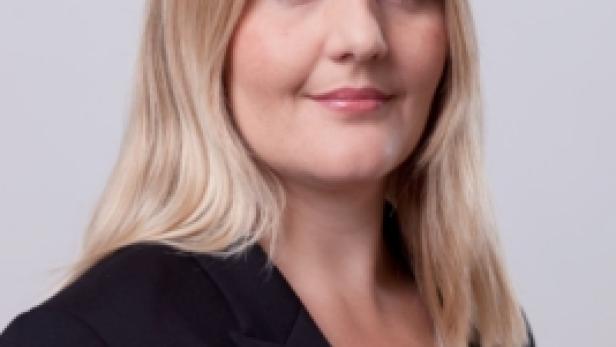 Eine Frau mit blonden Haaren und einem schwarzen Blazer lächelt in die Kamera.