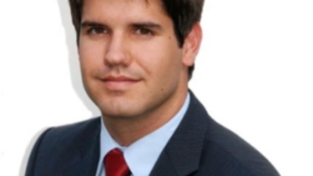 Ein Mann mit dunklen Haaren trägt einen dunklen Anzug mit roter Krawatte.