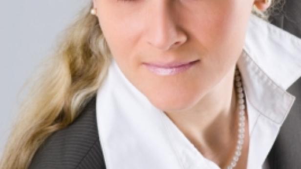 Eine blonde Frau in einem grauen Blazer und einer Perlenkette blickt in die Kamera.