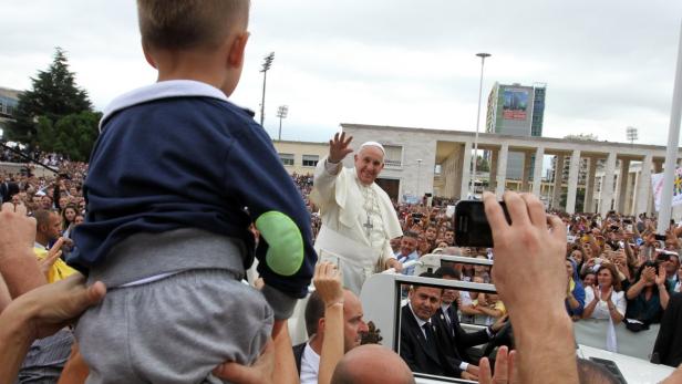 Papst Franziskus winkt einer Menschenmenge, während ein Kind auf den Schultern getragen wird.