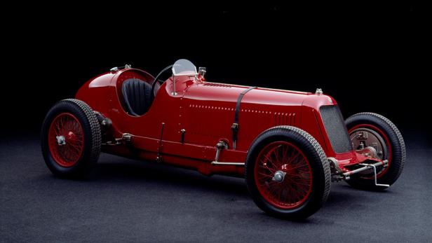 Ein roter Oldtimer-Rennwagen mit Speichenrädern vor schwarzem Hintergrund.