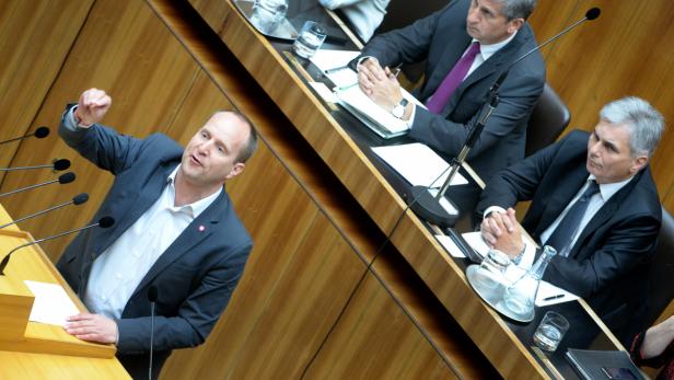 Ein Mann gestikuliert an einem Rednerpult während einer Sitzung, während andere Männer zuhören.