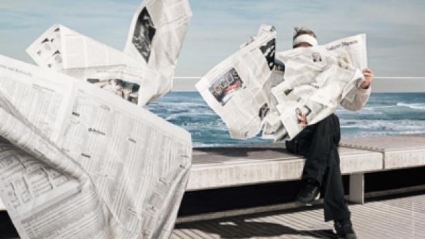 Ein Mann sitzt am Meer, umgeben von Zeitungen, die ihn fast vollständig bedecken.