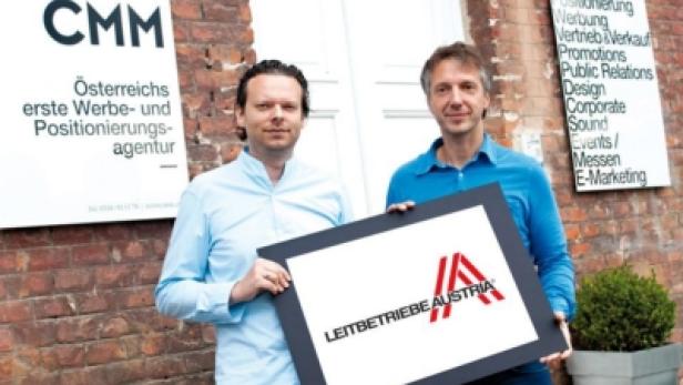 Zwei Männer präsentieren ein Schild mit dem Logo von „Leitbetriebe Austria“ vor einem Bürogebäude.