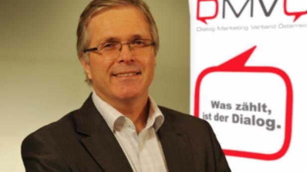 Ein Mann mit Brille und Anzug lächelt vor einem DMVÖ-Logo.