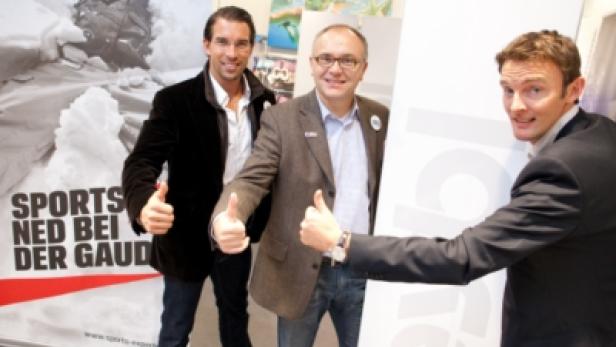 Drei Männer posieren lächelnd mit erhobenem Daumen vor einem Banner von „Sports Experts“.