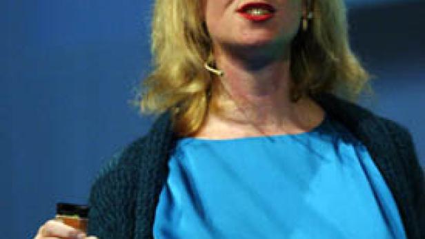 Eine blonde Frau in einem blauen Kleid hält ein Glas und spricht vor einem Publikum.