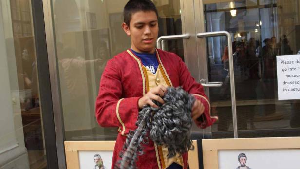 Ein junger Mann im historischen Kostüm hält eine graue Perücke in den Händen.