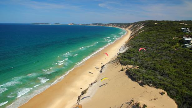 Ein Panoramablick auf einen langen Sandstrand mit türkisfarbenem Wasser und mehreren Gleitschirmfliegern.