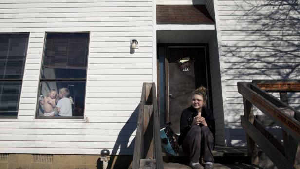 Eine junge Frau sitzt auf einer Treppe vor einem Haus, im Fenster sind zwei Kinder zu sehen.