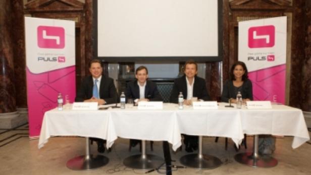 Vier Personen sitzen an einem Tisch mit „PULS 4“-Logos im Hintergrund.